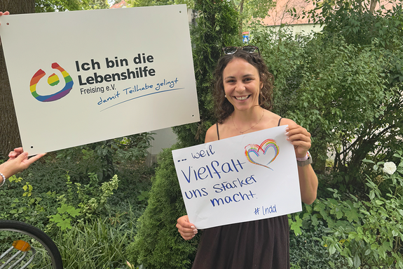 Alexandra Deml feiert die Demokratie weil Vielfalt uns stärker macht. Foto: Lebenshilfe Eine junge Frau mit Brille im Haar steht im Grünen und hält ein Schild, daneben hält jemand ein Schild "Ich bin die Lebenshilfe Freising e.V."