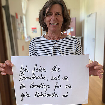 Claudia Nertinger feiert die Demokratie, weil sie die Grundlage für ein gutes Miteinander ist. Foto: Claudia Bauer Eine Frau mit halblangen Haaren und gestreiftem Oberteil steht in einem Gang und hält ein Schild.