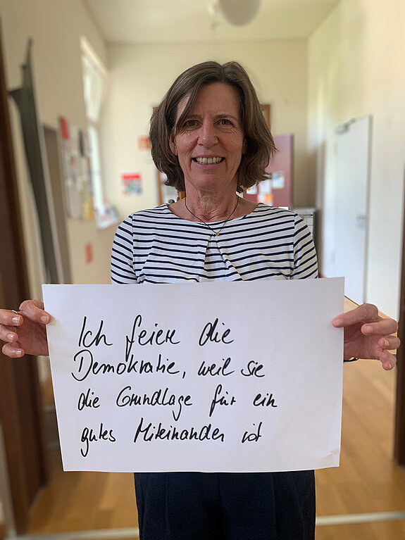 Claudia Nertinger feiert die Demokratie, weil sie die Grundlage für ein gutes Miteinander ist. Foto: Claudia Bauer Eine Frau mit halblangen Haaren und gestreiftem Oberteil steht in einem Gang und hält ein Schild.