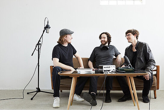 drei jung Männer auf einem Sofa, davor ein niedriger Tisch, daneben eine Stehlampe