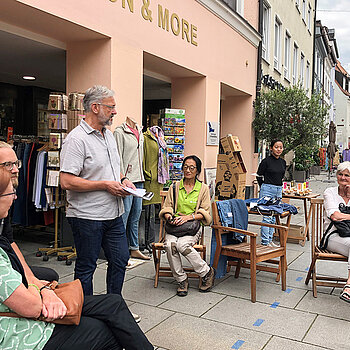 Mehrere Menschen sitzen vor einem Laden auf Stühlen und hören einem Mann zu. 