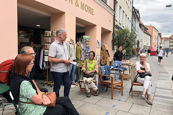 Mehrere Menschen sitzen vor einem Laden auf Stühlen und hören einem Mann zu. 