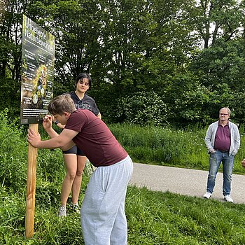 Ein Jugendlicher pinselt an einem Plakat im Grünen, beobachtet von zwei weiteren Personen