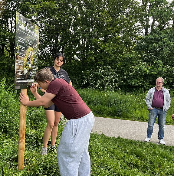 Ein Jugendlicher pinselt an einem Plakat im Grünen, beobachtet von zwei weiteren Personen