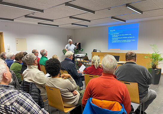 Publikum in Stuhlreihen von hinten, vorne eine Präsentation in blau und die Referentin