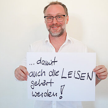 Stadtratsmitglied Reinhard Fiedler. Foto: Stadt Freising Ein Mann in einem weißen Hemd zeigt ein Schild mit der Aufschrift "damit auch die LEISEN gehört werden!"