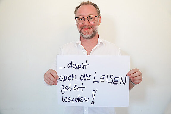 Stadtratsmitglied Reinhard Fiedler. Foto: Stadt Freising Ein Mann in einem weißen Hemd zeigt ein Schild mit der Aufschrift "damit auch die LEISEN gehört werden!"