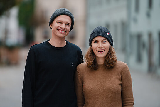 Junger Mann in dunklem Pulli mit Strickmütze, junge Frau im braunen Pulli mit Strickmütze, beide lachen