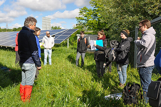 Professor Reinke mit Studierenden auf einer Freiflächenanlage. Foto: Markus Reinke Mehrere Menschen stehen im Kreis auf einer Wiese vor einer Freiflächen-PV Anlage