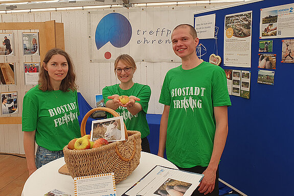 zwei Frauen und ein Mann in grünen T-Shirts mit Aufdruck "Biostadt Freising" stehen hinter einem Stehtisch mit Apfekorb darauf. Daneben eine Pinnwand mit Infos.