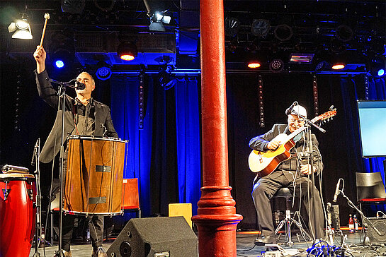 zwei Musiker mit Gitarre und Percussion auf der Bühne, der Referent auf der Videoleinwand im Hintergrund