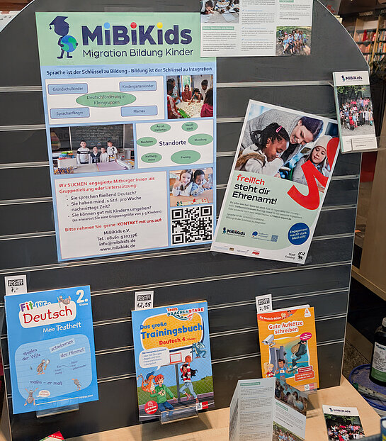 MiBiKids unterstützt Kinder beim Erlernen der deutschen Sprache. Foto: Stadt Freising Zwei Plakate und einige Deutsch-Trainingsbücher sind an einer grauen Rückwand angebracht.