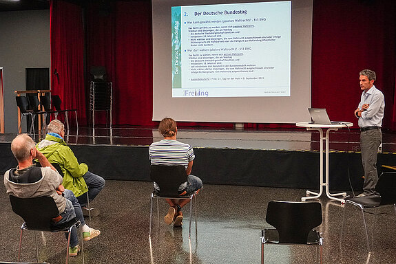 Michael Eberwein steht am Stehtisch, hinter ihm die Präsentation an der Wand und einige Zuhörer von hinten.