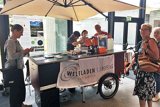 Mehrere Menschen stehen um einen hüfthohen Wagen mit Sonnenschirm und Weltladen-Transparent, auf dem Kaffe gekocht wird