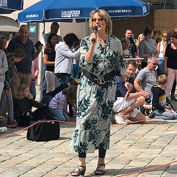 Frau im grün gemusterten Kleid mit Mikrofon und Sonnenbrille steht vor einer Menschenmenge und spricht.