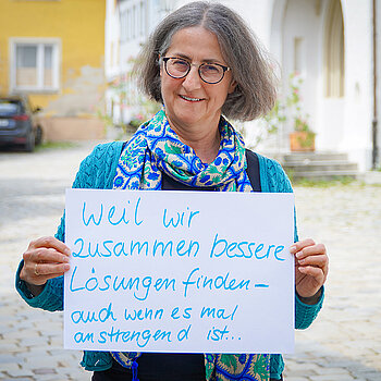 Johanna Sticksel (Treffpunkt Ehrenamt der Stadt Freising) feiert die Demokratie, weil wir zusammen bessere Lösungen finden - auch wenn es mal anstrengend ist. Foto: Stadt Freising Eine Frau mit Brille und halblangen Haaren steht im Freien und hält ein Schild. Im Hintergrund Kirchenportal, Baum und Auto.