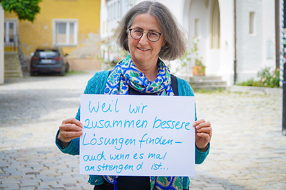 Johanna Sticksel (Treffpunkt Ehrenamt der Stadt Freising) feiert die Demokratie, weil wir zusammen bessere Lösungen finden - auch wenn es mal anstrengend ist. Foto: Stadt Freising Eine Frau mit Brille und halblangen Haaren steht im Freien und hält ein Schild. Im Hintergrund Kirchenportal, Baum und Auto.