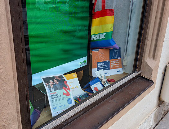 Engagement für den VdK, Fenster Marienapotheke. Foto: Stadt Freising vor grünem Hintergrund eine Regenbogen-Tasche "VdK", ein Plakat und Broschüren.