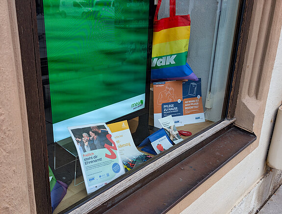 Engagement für den VdK, Fenster Marienapotheke. Foto: Stadt Freising vor grünem Hintergrund eine Regenbogen-Tasche "VdK", ein Plakat und Broschüren.