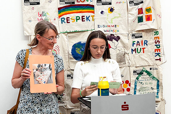 ein junges Mädchen und eine Frau vor einer Wand mit bedruckten Taschen 