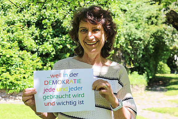 Marina Freudenstein hält ein Schild mit bunten Buchstaben: "... weil in der Demokratie jede und jeder gebraucht wird und wichtig ist."
