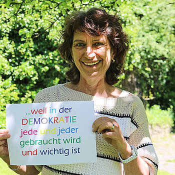 Marina Freudenstein hält ein Schild mit bunten Buchstaben: "... weil in der Demokratie jede und jeder gebraucht wird und wichtig ist."