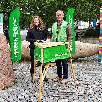 ein Mann und eine junge Frau neben einem grünen Tisch mit grünen Greenpeace-Flaggen im Hintergrund.