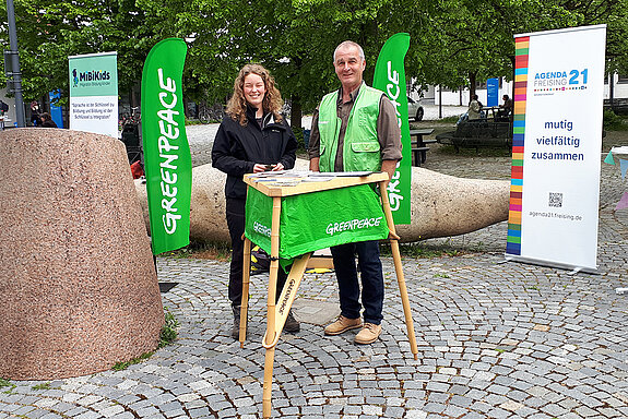 ein Mann und eine junge Frau neben einem grünen Tisch mit grünen Greenpeace-Flaggen im Hintergrund.