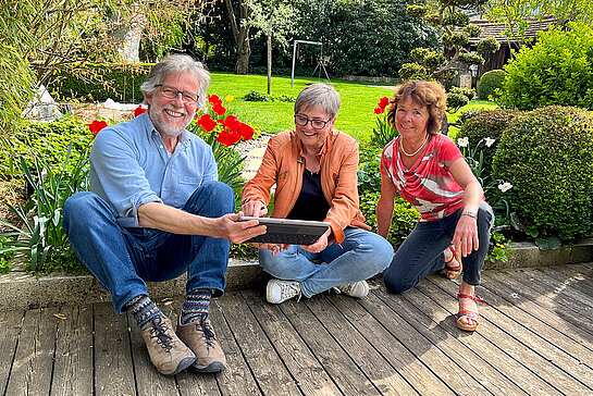 ein Mann und zwei Frauen mit Laptop auf einer Holzterasse am Boden sitzend, hinter sich den grünen Garten