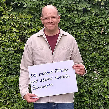 Oliver Sturde feiert die Demokraktie, weil sie Frieden sichert und soziale Bindungen stärkt. Foto: Feuerwehr Freising Ein Mann in heller Jacke steht vor einer Hecke und hält ein Schild.
