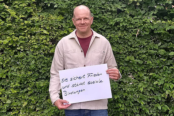 Oliver Sturde feiert die Demokraktie, weil sie Frieden sichert und soziale Bindungen stärkt. Foto: Feuerwehr Freising Ein Mann in heller Jacke steht vor einer Hecke und hält ein Schild.