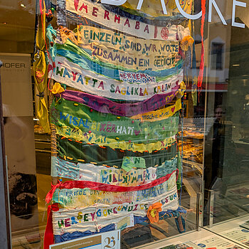 ein bunter Teppich aus Stoffstreifen in verschiedenen Sprachen. "Einzeln sind wir Worte, zusammen sind wir ein Gedicht."