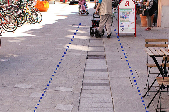 Pflasterausschnitt, bei dem der Leitstreifen frei bleibt. Daneben sind Tisch und Stühle sowie Aufsteller und weiter hinten Fahrräder zu sehen. Eine Person mit Rollator nutzt den freien Bereich.