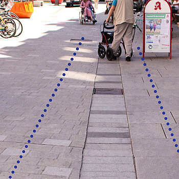 Pflasterausschnitt, bei dem der Leitstreifen frei bleibt. Daneben sind Tisch und Stühle sowie Aufsteller und weiter hinten Fahrräder zu sehen. Eine Person mit Rollator nutzt den freien Bereich.