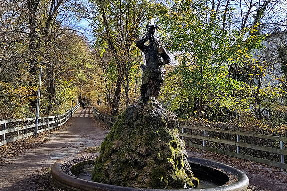 Im Vordergrund ein Brunnen, der Weg führt rechts und links gesäumt von herbstbunten Bäumen geradeaus