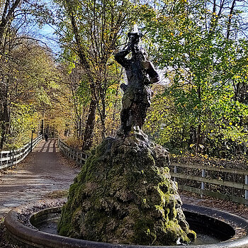 Im Vordergrund ein Brunnen, der Weg führt rechts und links gesäumt von herbstbunten Bäumen geradeaus