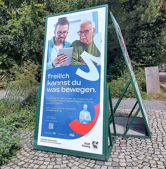 freilich kannst Du was bewegen... zwischen den Generationen. Foto: Stadt Freising ein übermannsgroßer Aufsteller auf Kopfsteinpflaster und mit Bäumen im Hintergrund. Vorwiegend in blautönen und mit einem jüngeren und einem älteren Mann, die sich unterhalten.