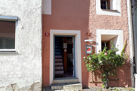 Hausfassade in Altrosa mit geöffneter Tür. Daneben ein Rosenstrauch
