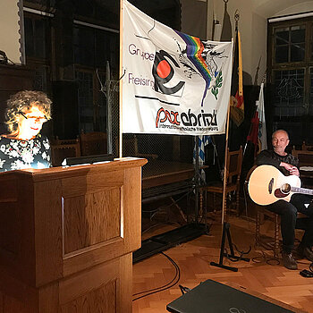 Marina Freudenstein am Stehpult, vor dem Pax-Christi-Transparent ein Gitarrist.