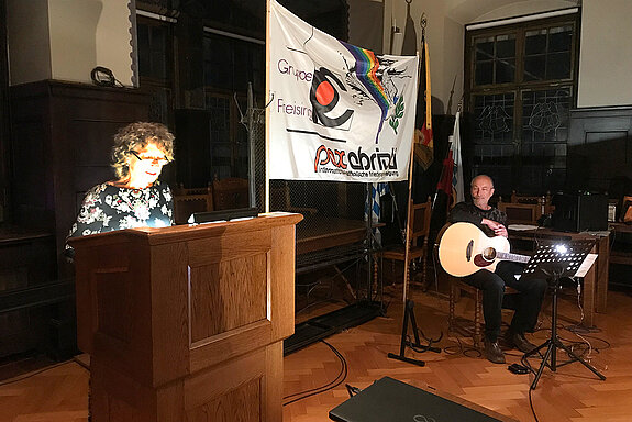 Marina Freudenstein am Stehpult, vor dem Pax-Christi-Transparent ein Gitarrist.