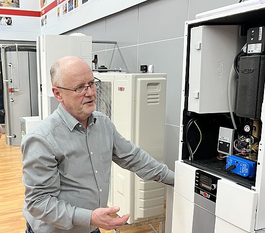 Ein Mann mit Brille und im grauen Hemd steht erklärend vor einer geöffneten Wärmepumpen. Im Hintergrund stehen weitere 