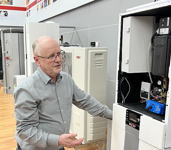 Ein Mann mit Brille und im grauen Hemd steht erklärend vor einer geöffneten Wärmepumpen. Im Hintergrund stehen weitere 