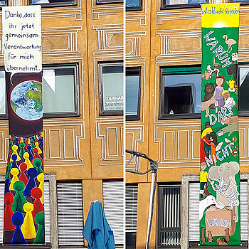 7 Meter lange bunt bemalte Banner an der Hausfassade. Eine Erde mit Sprechblase, darunter Mensch-ärgere-dich-Männchen. Auf dem anderen Banner viele Tiere und Menschen, die wählen wollen 