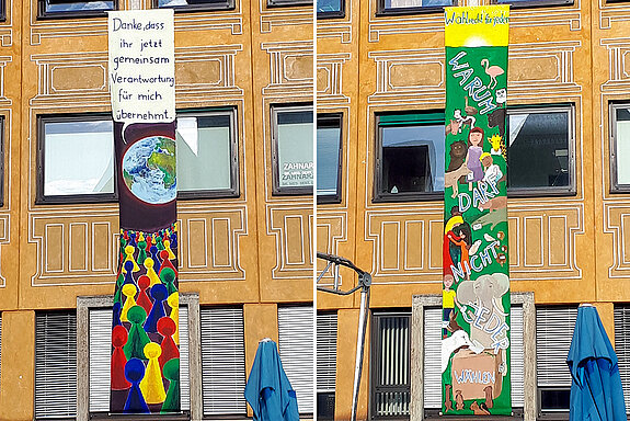 7 Meter lange bunt bemalte Banner an der Hausfassade. Eine Erde mit Sprechblase, darunter Mensch-ärgere-dich-Männchen. Auf dem anderen Banner viele Tiere und Menschen, die wählen wollen 