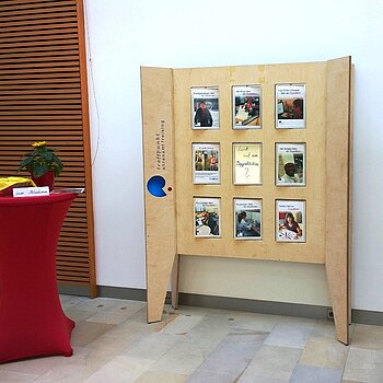 Ausstellungswand aus Holz mit beweglichen Elementen. Auf diesen sind Fotos und Texte zu Menschen, die sich engagieren. In der Mitte ein Spiegel mit der Aufschrift: "Lust auf ein Doppelleben?"