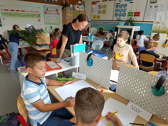 Kinder sitzen in einem Klassenzimmer und malen. Eine Lehrerin steht an einem Tisch und schaut ihnen dabei zu.