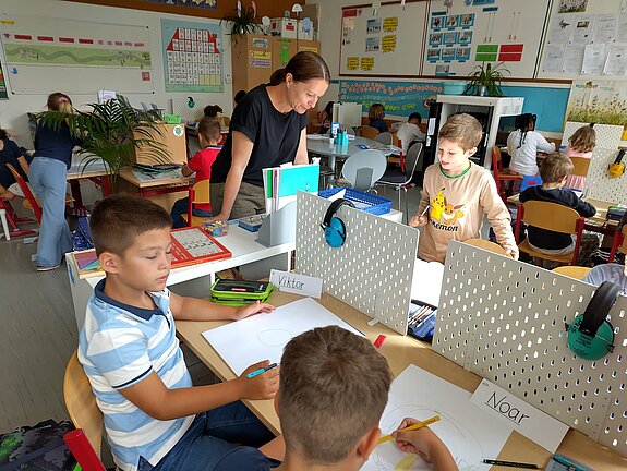 Kinder sitzen in einem Klassenzimmer und malen. Eine Lehrerin steht an einem Tisch und schaut ihnen dabei zu.