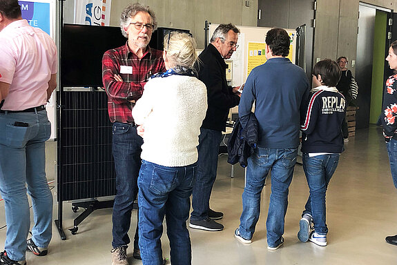 Zwei Männer beraten drei Personen. Im Hintergrund das schwarze Balkonsolarmodul.