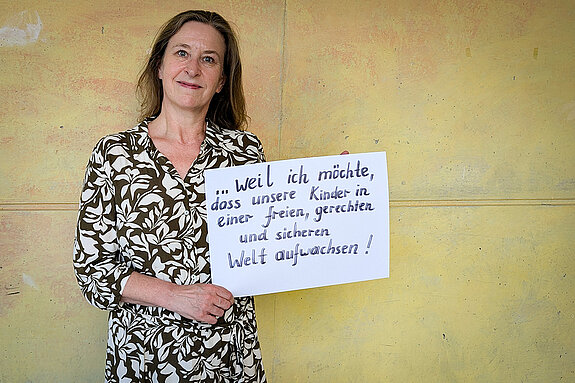 Gisela Schauz, Lehrerin am OMG feiert die Demokratie, weil sie möchte, dass unsere Kinder in einer freien, gerechten und sicheren Welt aufwachsen! dass unsere Kinder Eine Frau im weiß-braun gemusterten Kleid steht vor einer gelben Wand und hält ein Schild