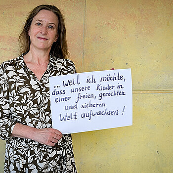 Gisela Schauz, Lehrerin am OMG feiert die Demokratie, weil sie möchte, dass unsere Kinder in einer freien, gerechten und sicheren Welt aufwachsen! dass unsere Kinder Eine Frau im weiß-braun gemusterten Kleid steht vor einer gelben Wand und hält ein Schild